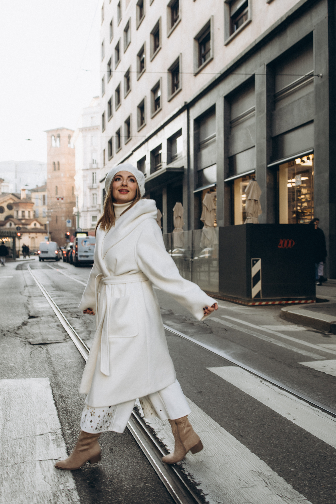 Winter Street Portrait on Milan's Tram Tracks A woman in a white coat and beret walks across tram tracks on a Milan street with a bell tower behind her in winter