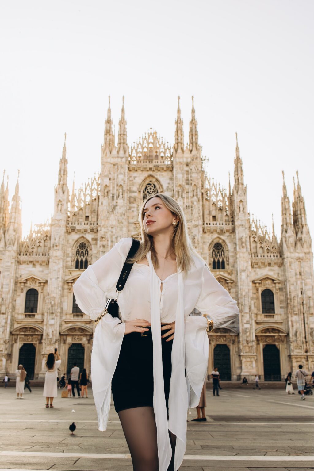 Woman in a white blouse and black skirt glancing up toward the spires of Milan Cathedral in soft backlight on Piazza del Duomo