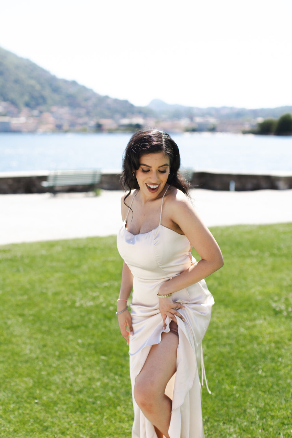 Woman in a champagne dress laughing and gathering her skirt on a lakeside lawn on Lake Como, the water and town behind her