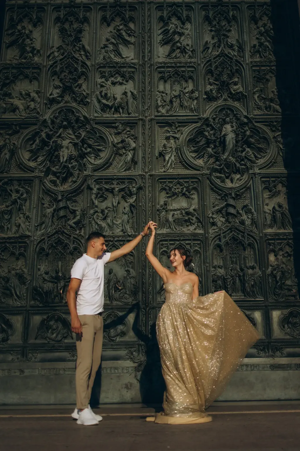 A man twirls a woman in a glittering gold gown in front of the carved bronze doors of the Duomo di Milano.