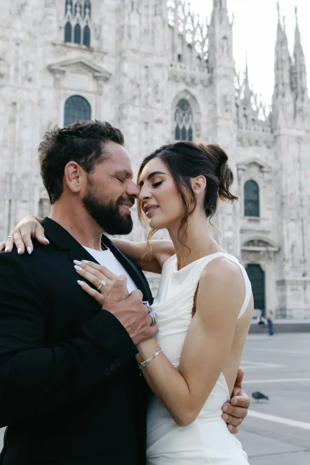 Close portrait of the couple cheek to cheek, his ringed hand on her arm and her eyes closed, before the white Duomo facade.
