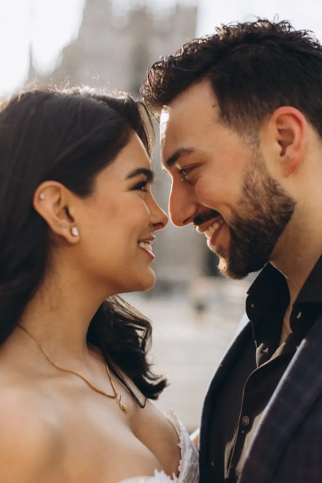 A warm close-up of the couple nose to nose and laughing, her pearl earring and gold necklace bright against the blurred Duomo.