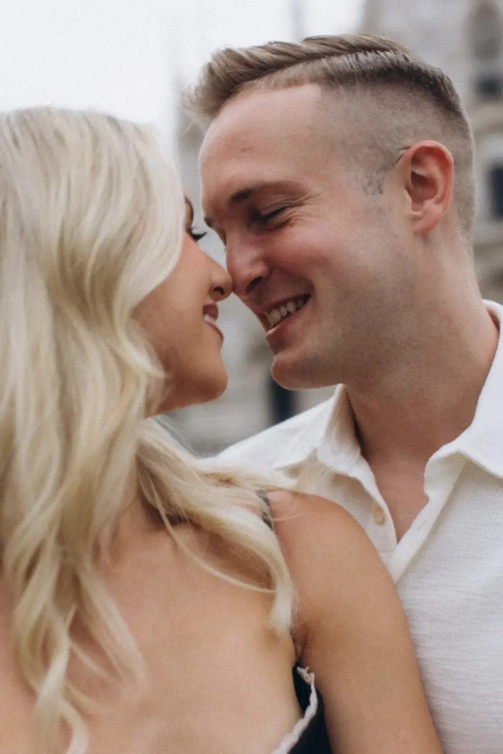 A tight close-up of the couple smiling nose to nose, her blonde waves in soft focus and the cathedral pale behind them.