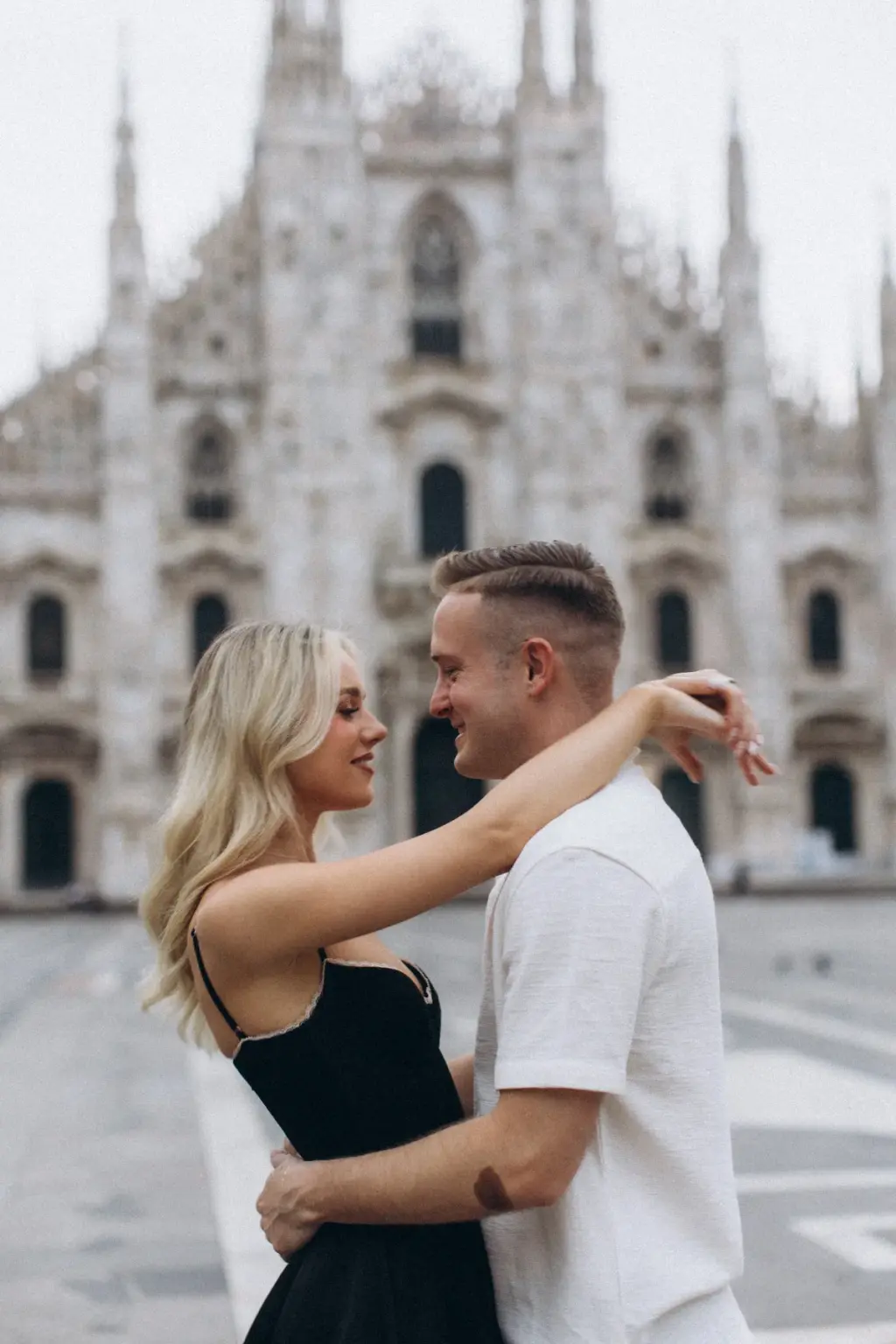 A woman in a black dress loops her arms around the neck of her partner in a white shirt, both laughing before the Duomo.