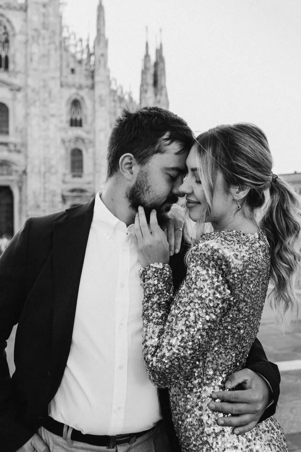 In black and white, a bearded man and a woman with a high ponytail touch foreheads, her hand on his jaw, by the Duomo spires.