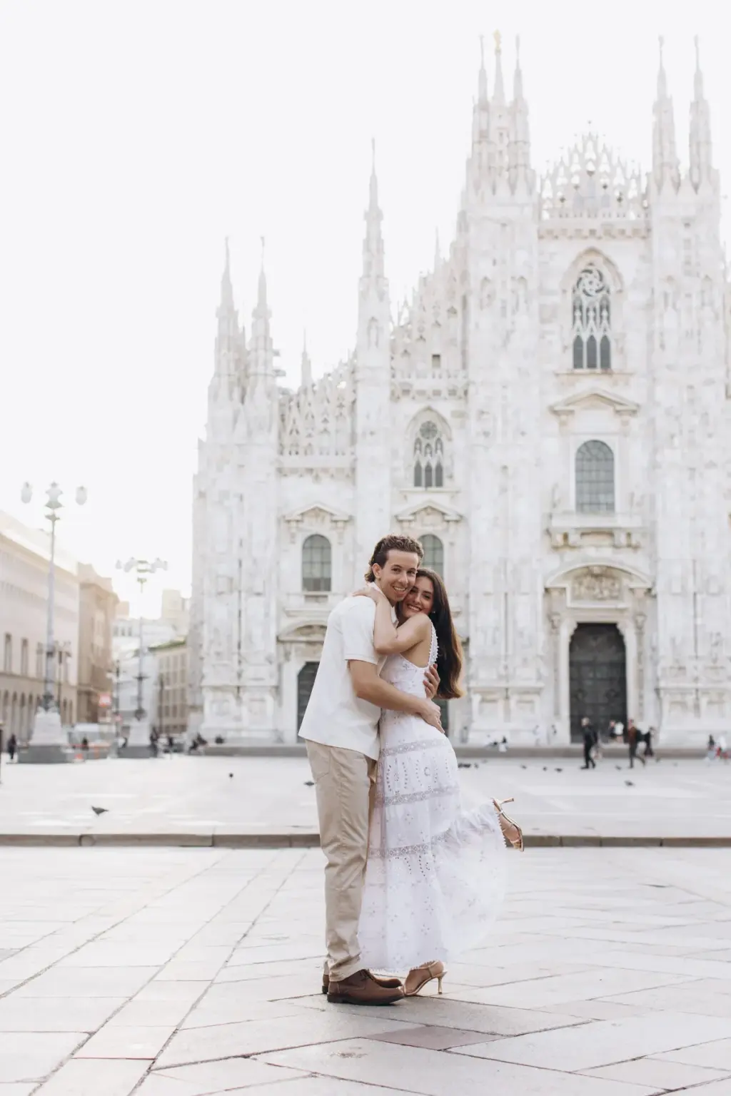 A woman in a white lace sundress hugs her partner in a white polo in the near-empty Piazza del Duomo, the Duomo facade towering behind.