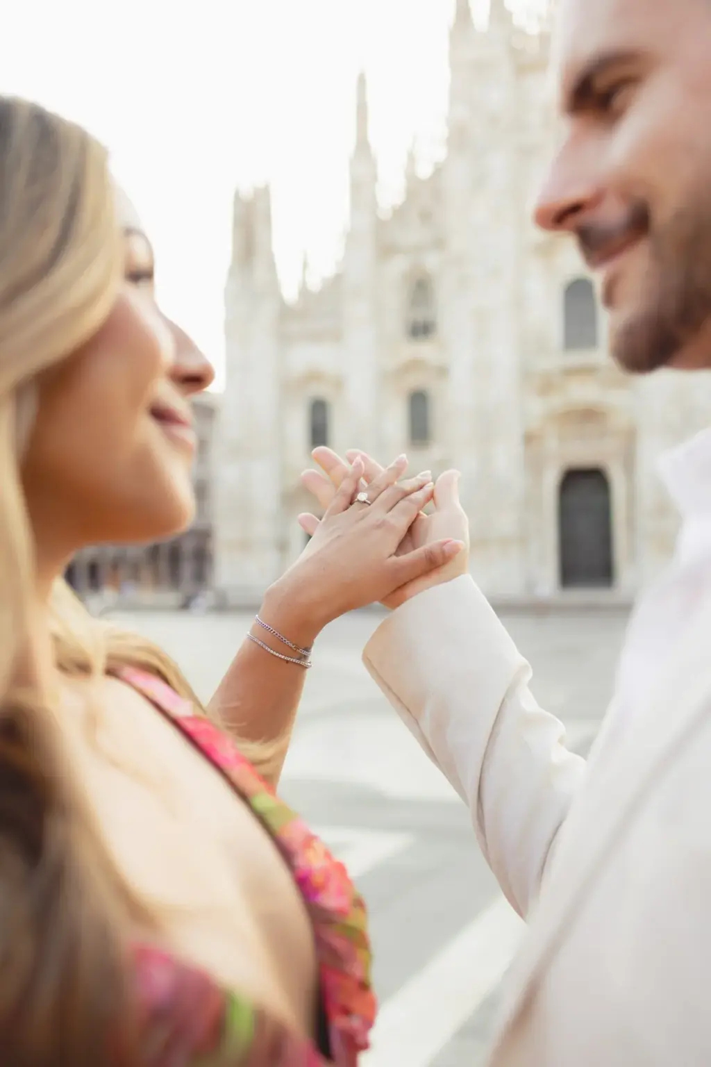 Close-up of their joined hands showing her engagement ring, both smiling, the Duomo facade blurred into the background.