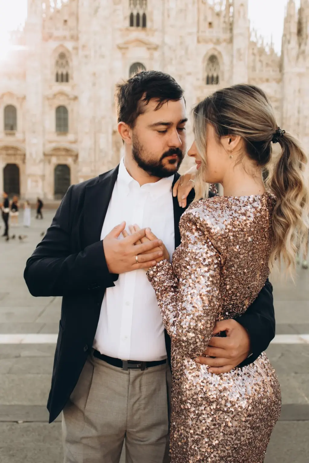 A bearded man in a blazer holds his partner's hand to his chest as she leans in, her sequin dress shimmering before the Duomo.