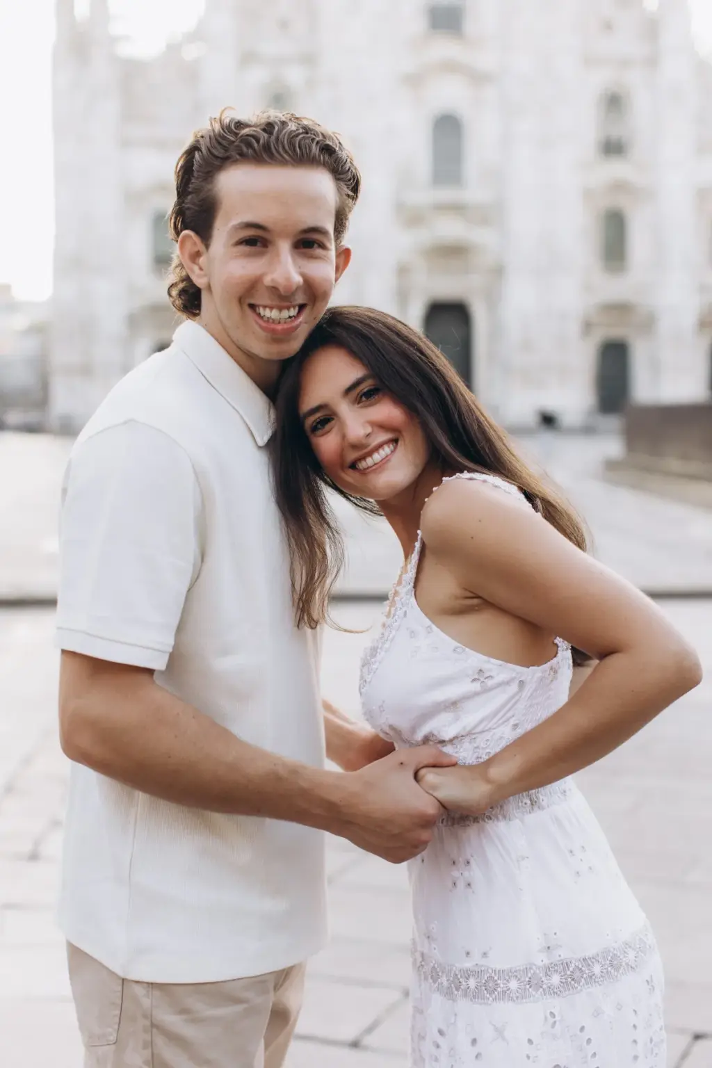 A laughing woman in a white lace dress leans her head on her partner's shoulder as they hold hands in front of the Duomo.