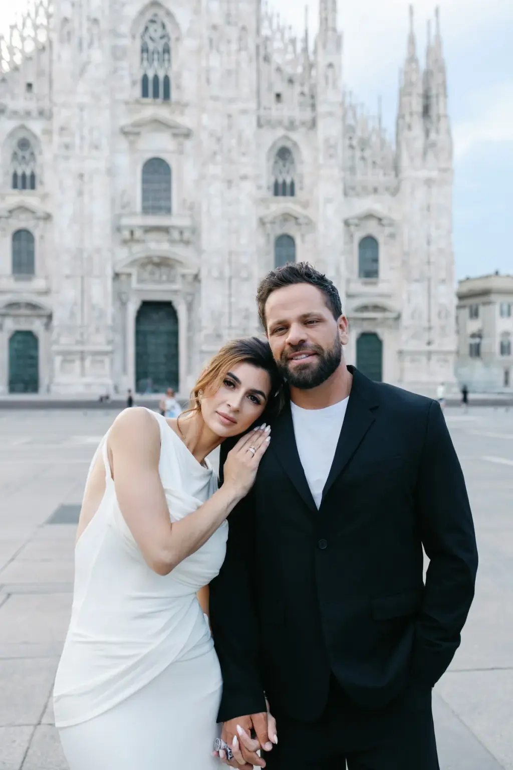 A woman in a white satin dress rests her head on her partner's shoulder in front of the Duomo di Milano facade.