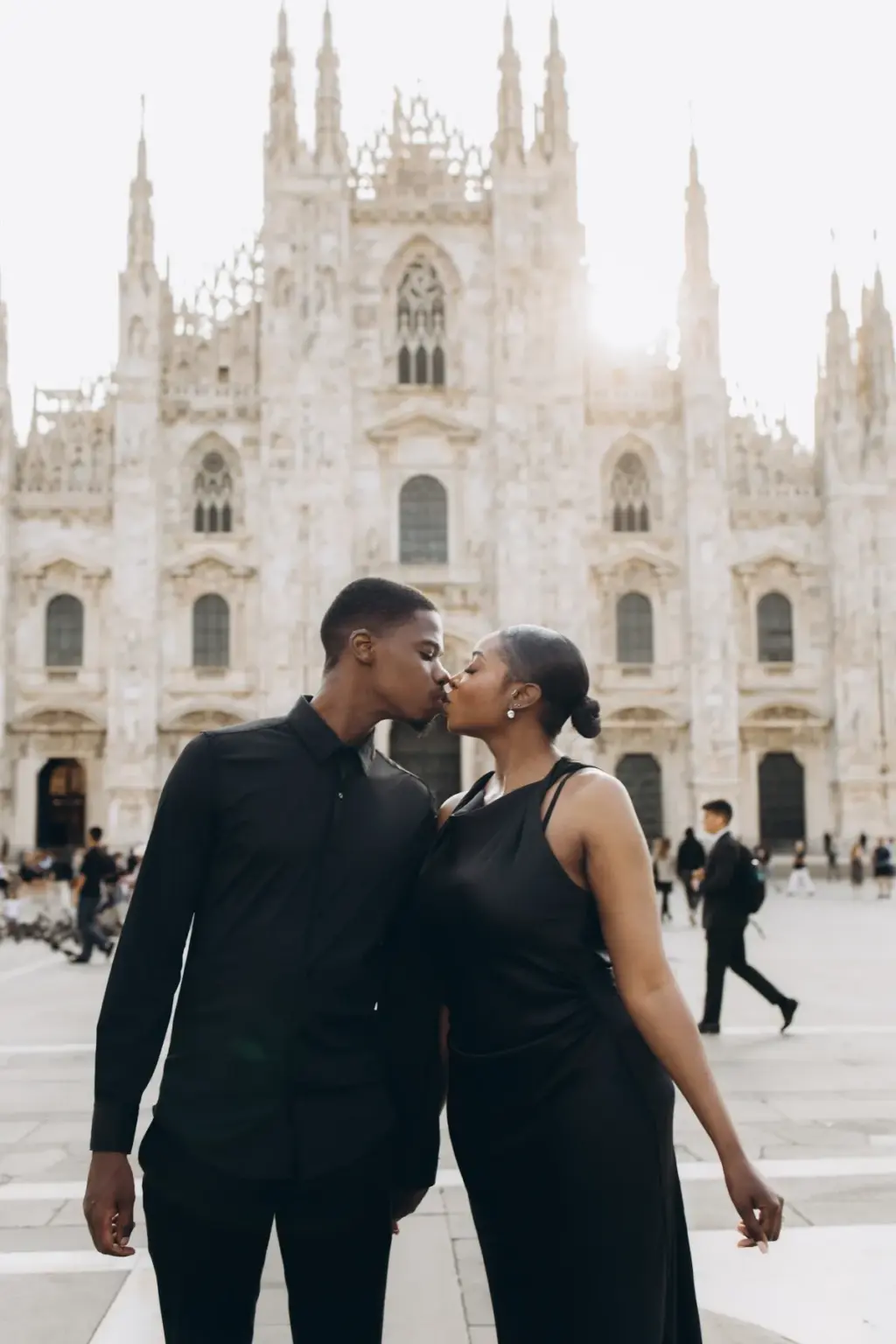 The couple leans in for a kiss on the Duomo square, both in black, with the white cathedral facade filling the frame behind.