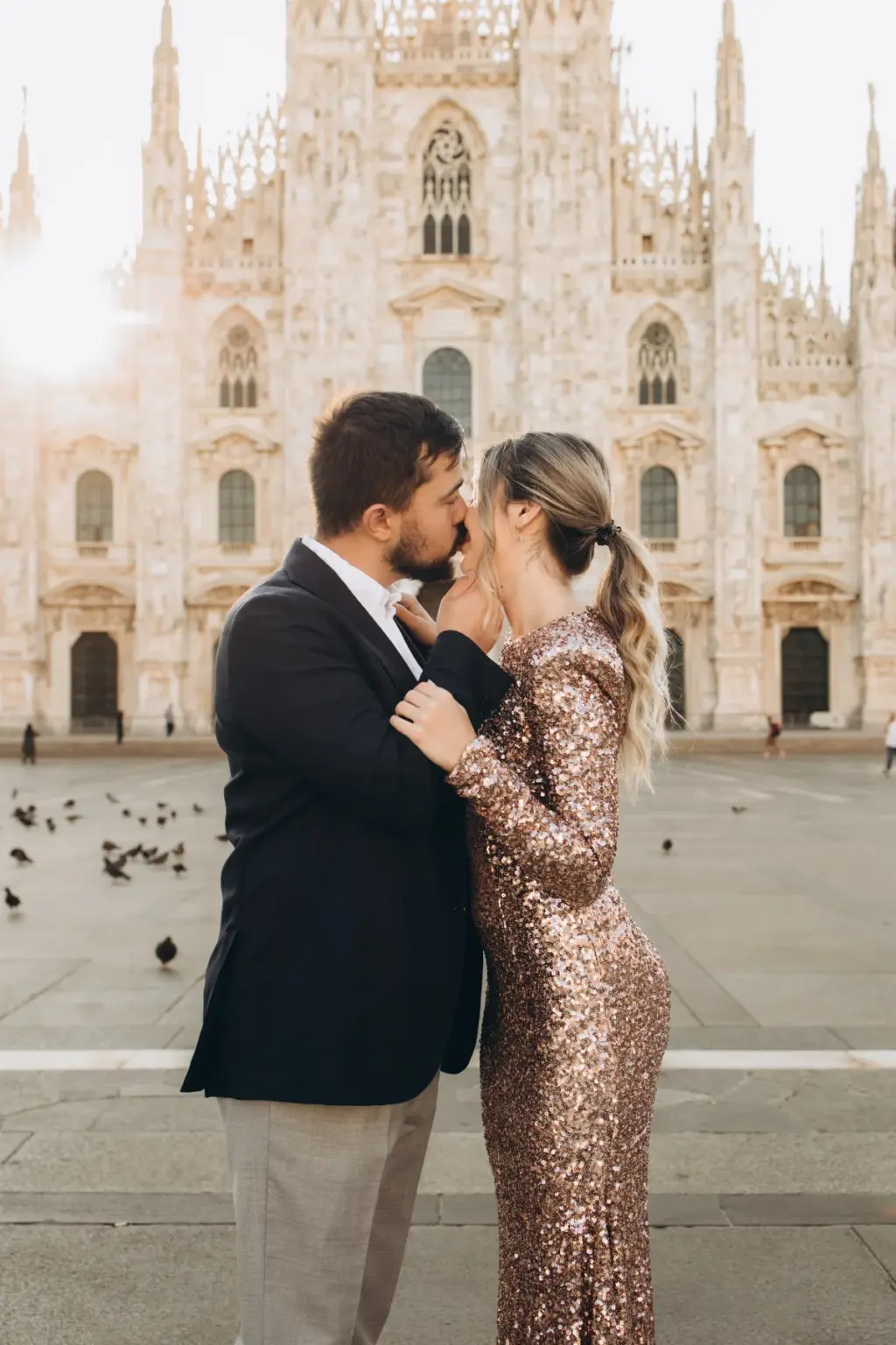 A couple in a sequin gown and dark blazer share a kiss in front of the Duomo while pigeons gather on the square around them.