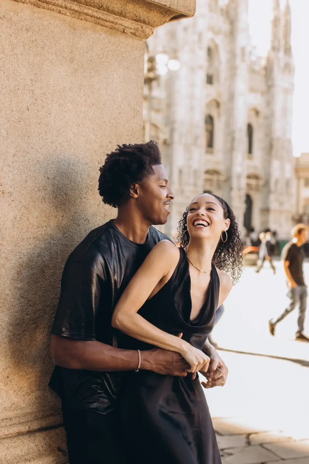 A woman in a black satin dress laughs out loud as her partner hugs her from behind by a stone pillar with the Duomo behind them.