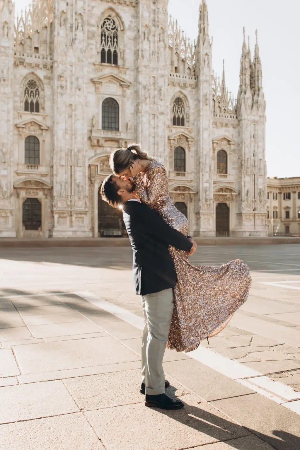 A man lifts a woman in a sequin gown off the ground, her skirt flaring as they kiss, on the open square in front of the Duomo.