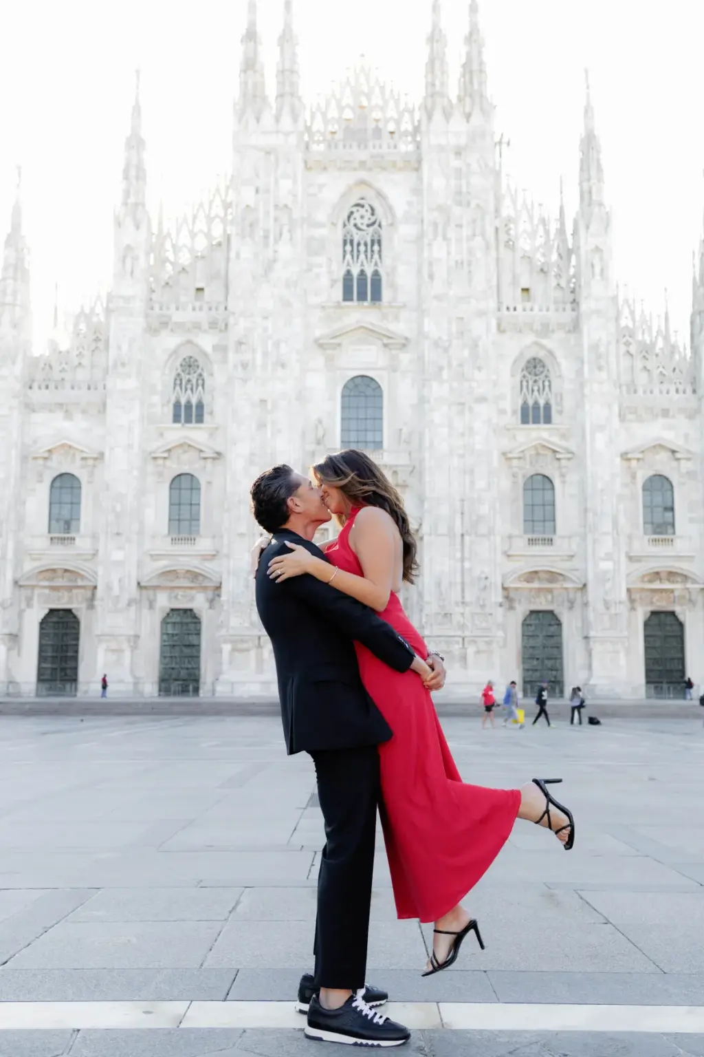 On the open Duomo square he lifts her off the ground mid-kiss, her red dress flowing and heels kicked up behind her.