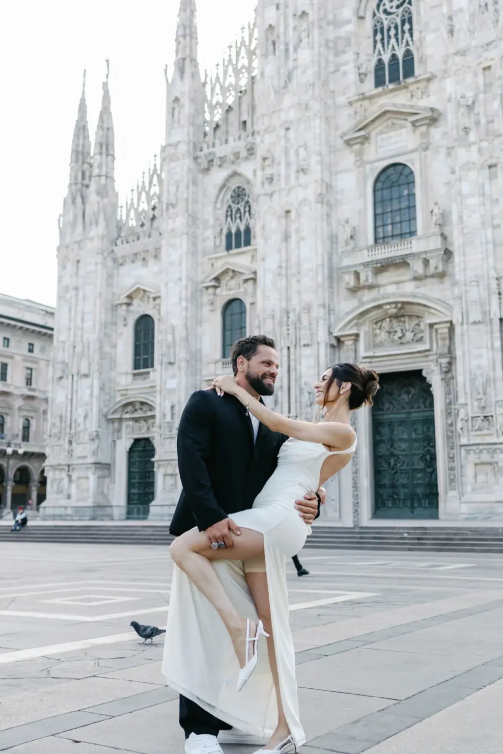 A bearded man lifts his partner in a white slit dress off the ground as she laughs, beside the towering Duomo di Milano.