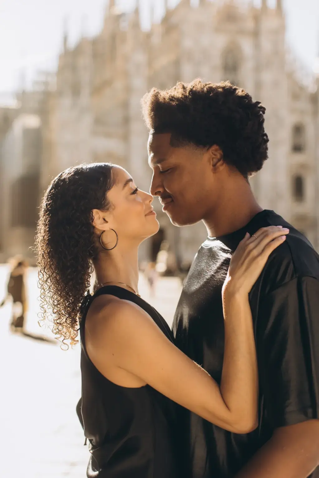 A couple in black stand nose to nose in front of the Duomo di Milano, her hand resting on his shoulder as they lean in close.
