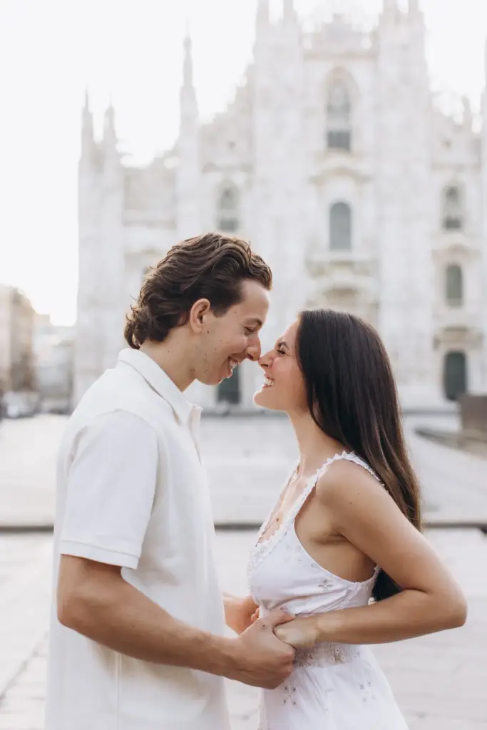 Nose to nose and smiling, a couple in white hold hands in front of the Duomo di Milano, its spires blurred in the background.