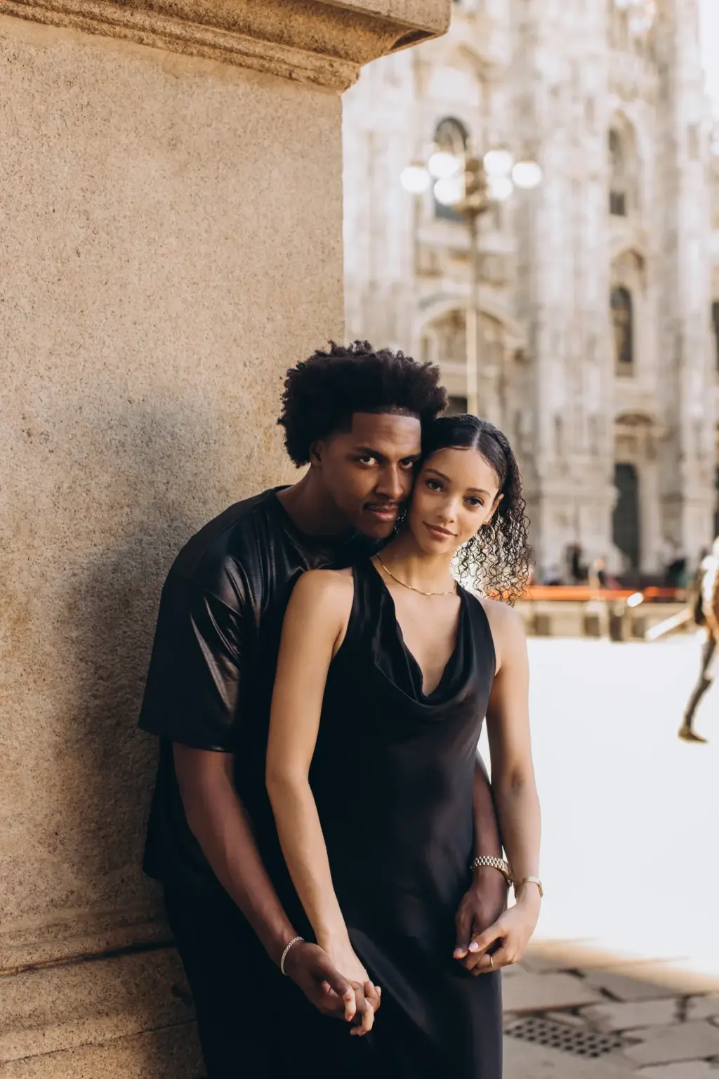 A man holds his partner from behind against a stone pillar, the Duomo facade and a lamppost rising behind them in Piazza del Duomo.