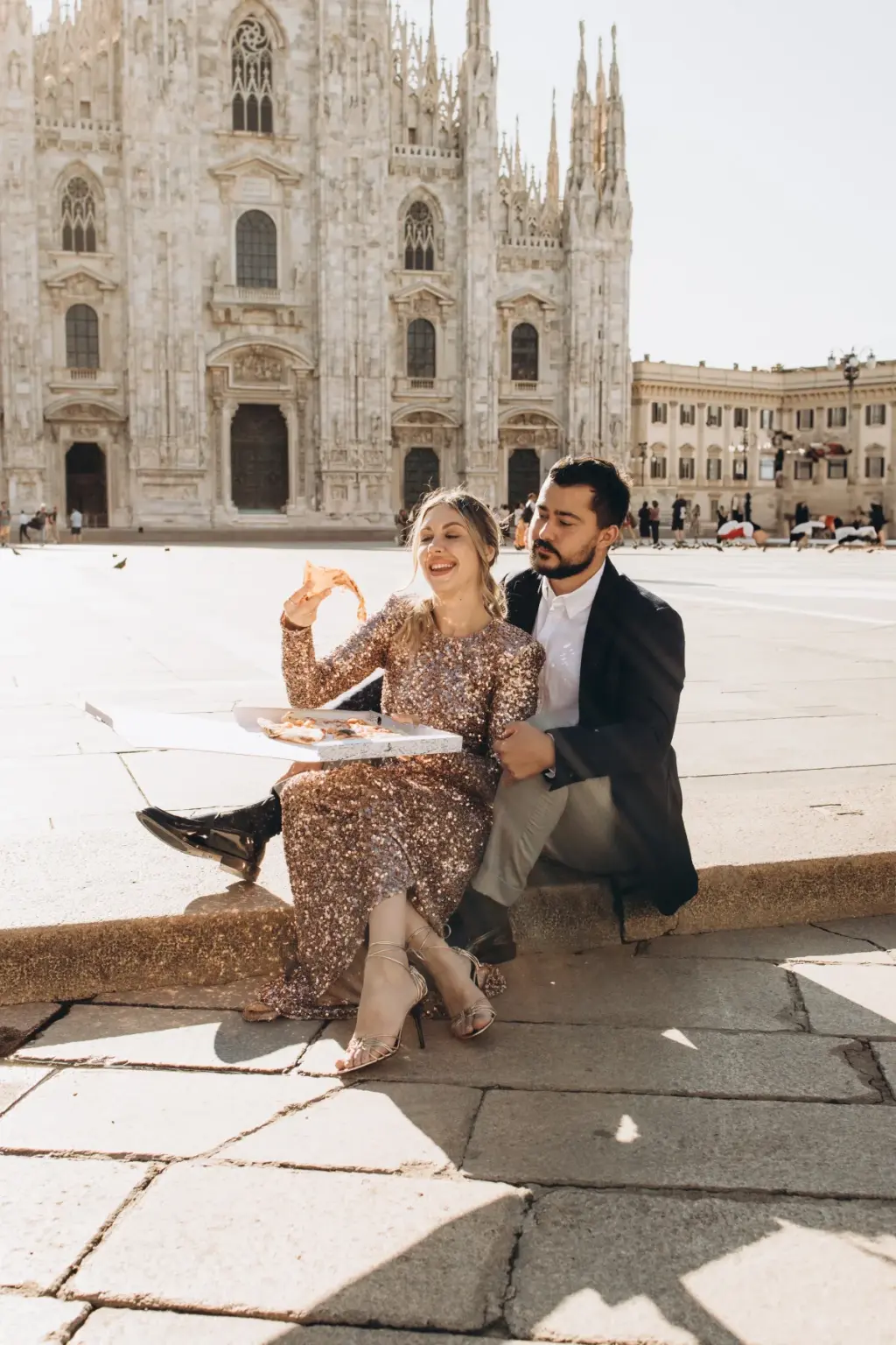 Sitting on a stone kerb by the Duomo, a laughing woman in a sequin gown holds up a slice of pizza beside her partner in a blazer.
