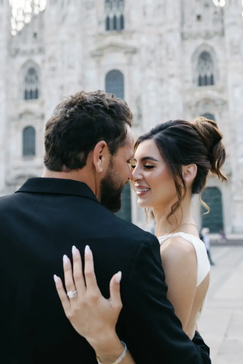 Her hand with an engagement ring rests on his black jacket as the couple leans in close, the Duomo facade softly blurred behind.