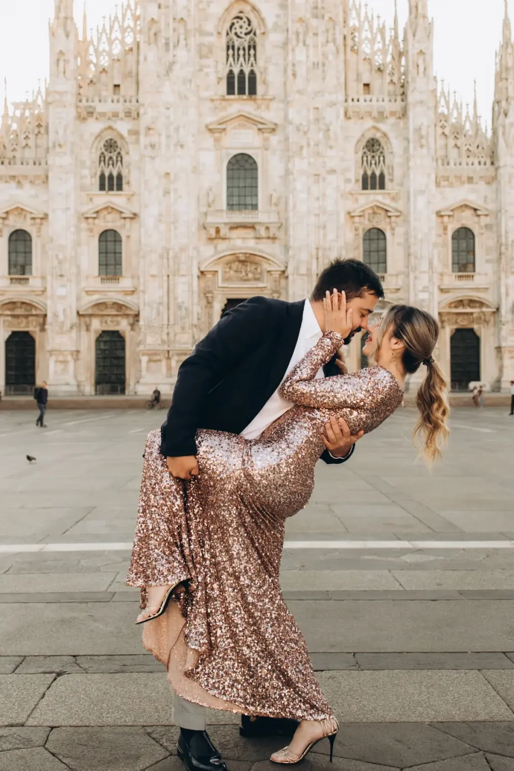 A man in a dark blazer dips a woman in a rose-gold sequin gown into a kiss in the middle of the square before the Duomo di Milano.