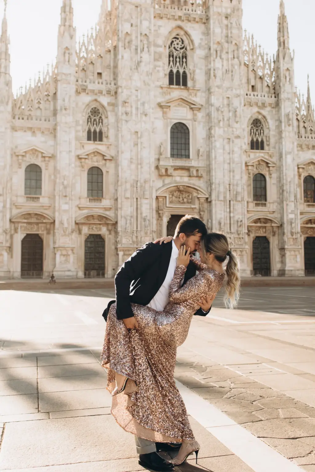 A man pulls a woman in a long sparkling gown into a deep embrace on the bright paved square below the white Duomo facade.