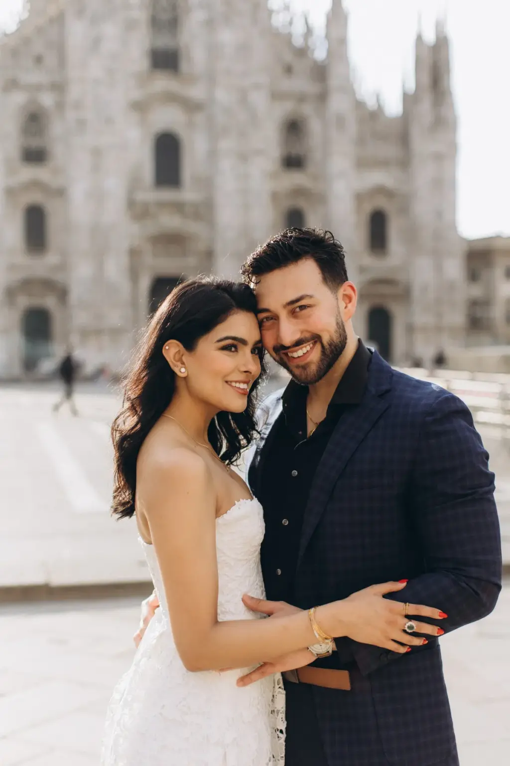 Both smiling into the camera, the couple stands cheek to cheek in front of the Duomo di Milano facade, her hand on his chest.