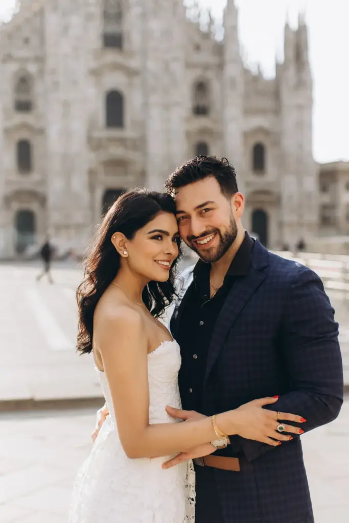 Both smiling into the camera, the couple stands cheek to cheek in front of the Duomo di Milano facade, her hand on his chest.