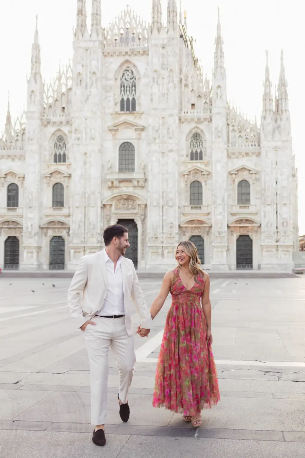 A man in a cream suit and a woman in a pink floral maxi dress walk hand in hand across the empty square before the Duomo.