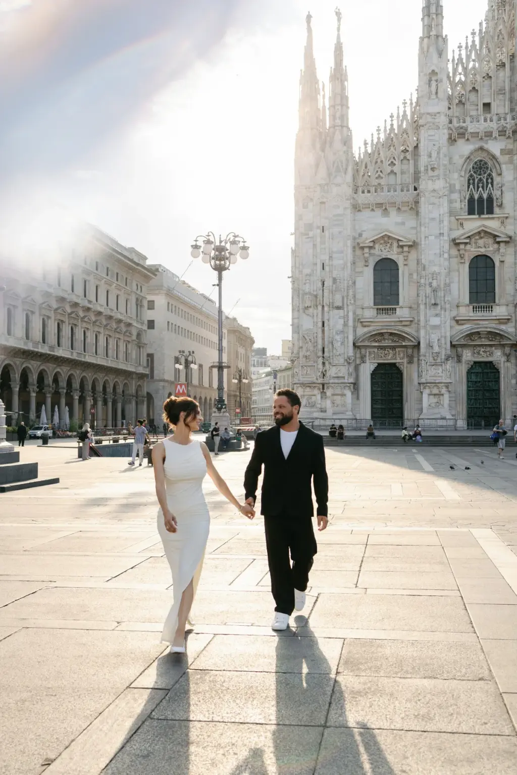 Hand in hand, the couple walks across the sunlit square toward the camera, the Duomo di Milano spires on their right.