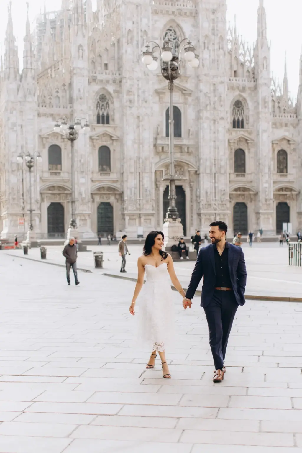A couple walks hand in hand toward the camera across the Duomo square, she in a white strapless dress and he in a navy suit.