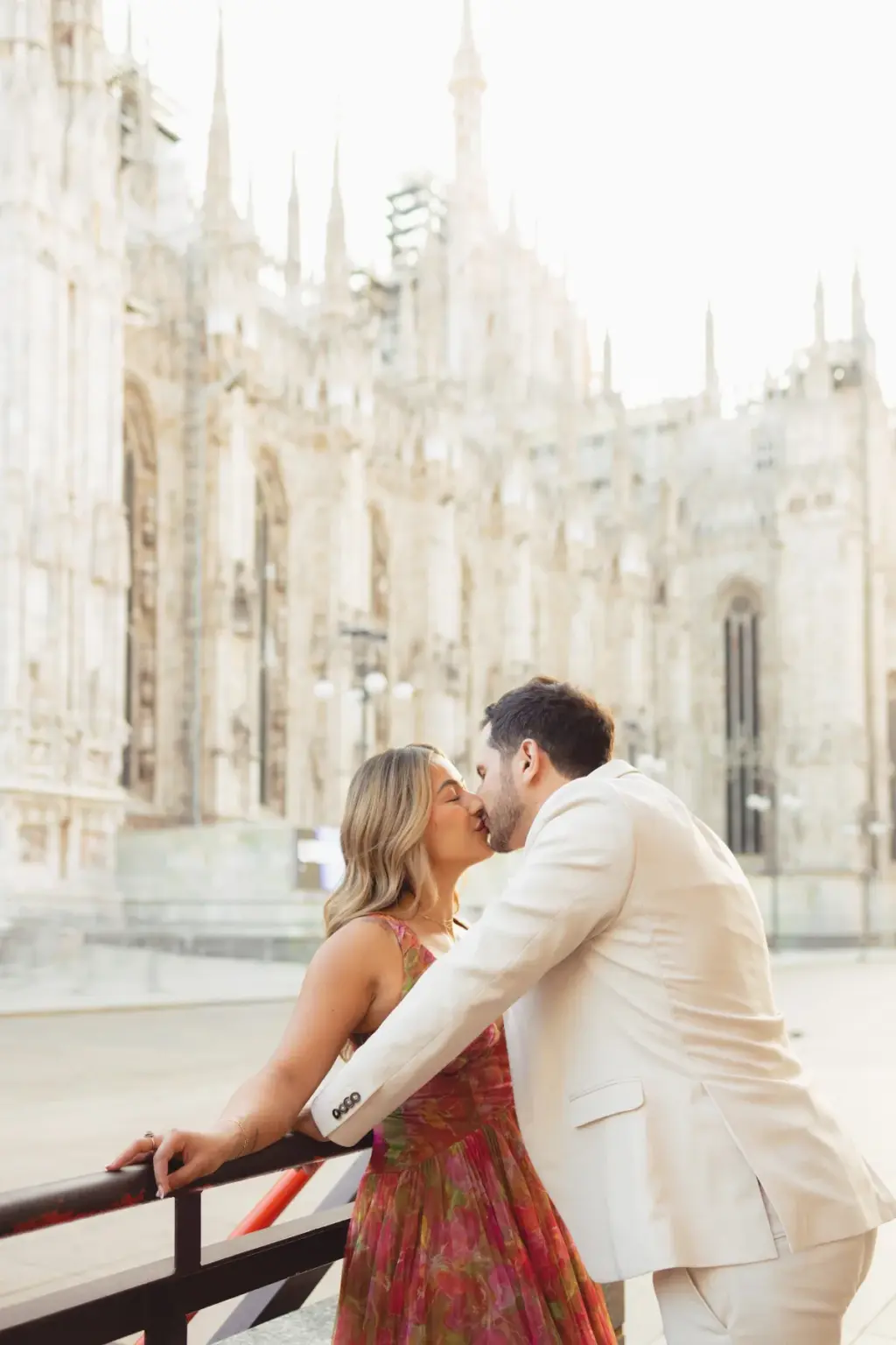 Leaning on a dark railing, she kisses him under the carved side walls and spires of the Duomo di Milano.