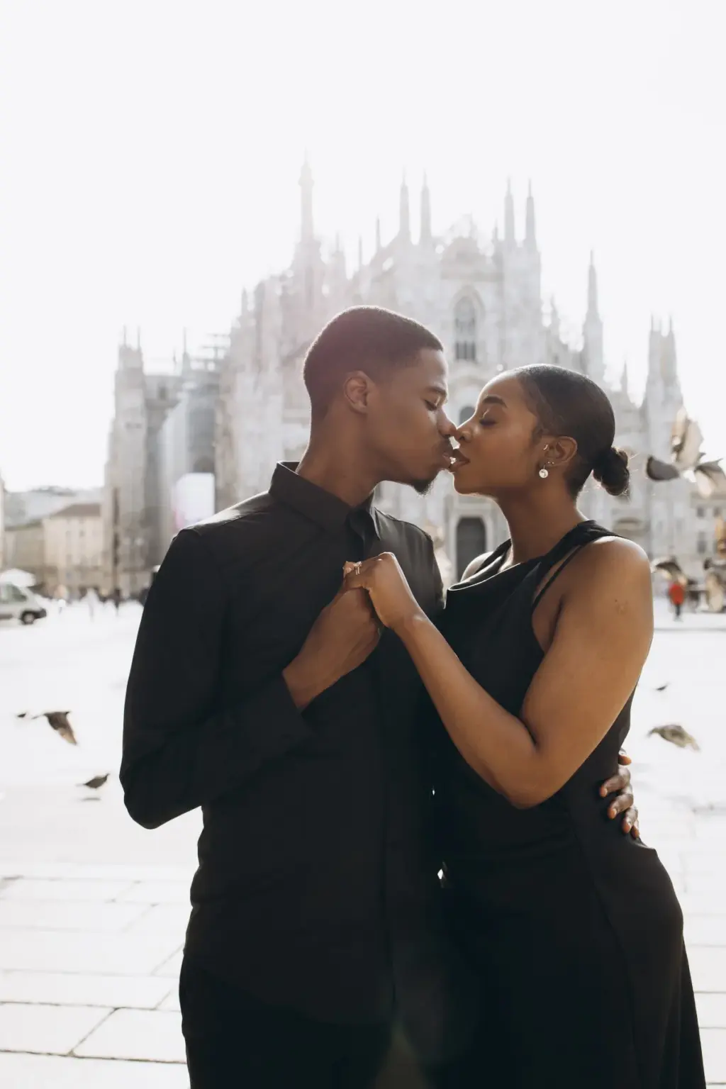 Almost kissing with hands joined between them, the couple stands on the Duomo square as pigeons flutter past the spires.