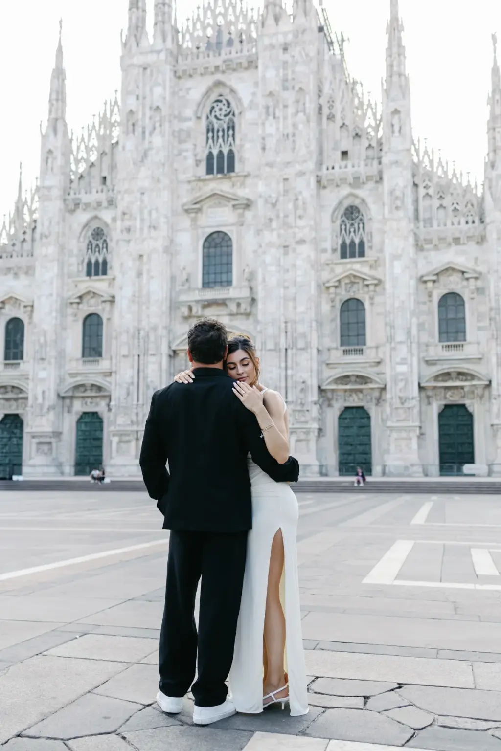 Seen from behind, a couple in black and white embraces on the empty Duomo square, facing the cathedral's gothic spires.