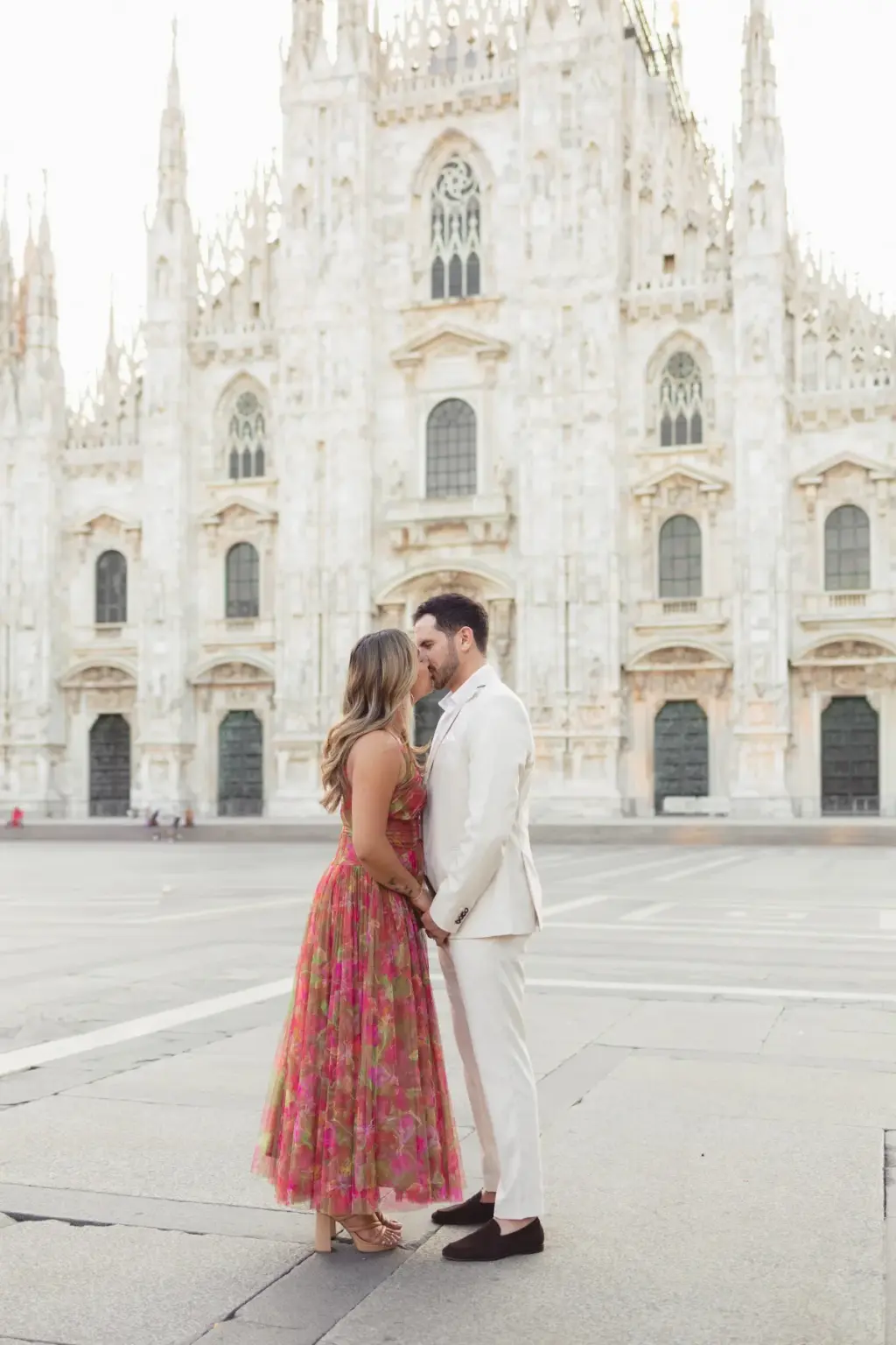 Full-length view of the couple standing close as he kisses her forehead, small figures against the Duomo's white front.