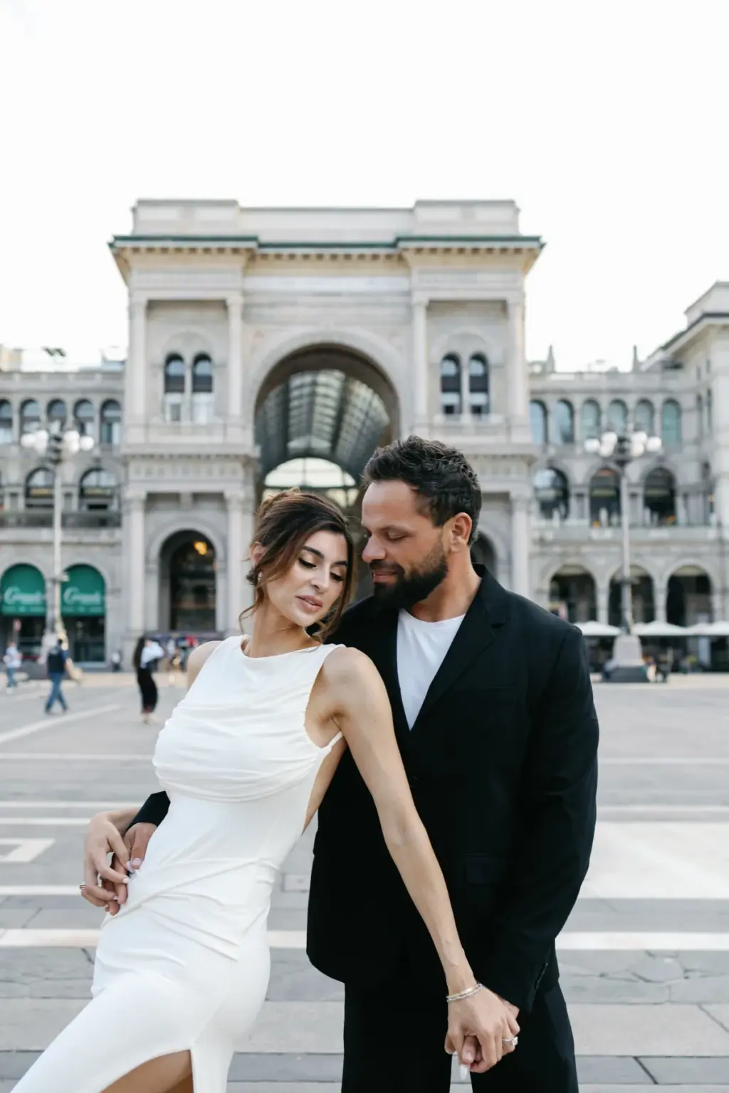 A woman in a white sleeveless dress leans back against her partner, hands linked, with the Galleria arch rising behind them.