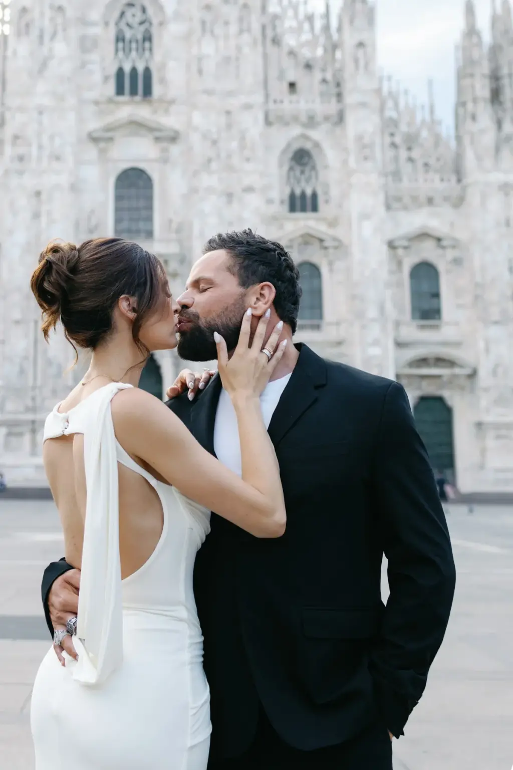 A couple shares a kiss on the Duomo square, her hand on his bearded cheek and the open back of her white dress in view.