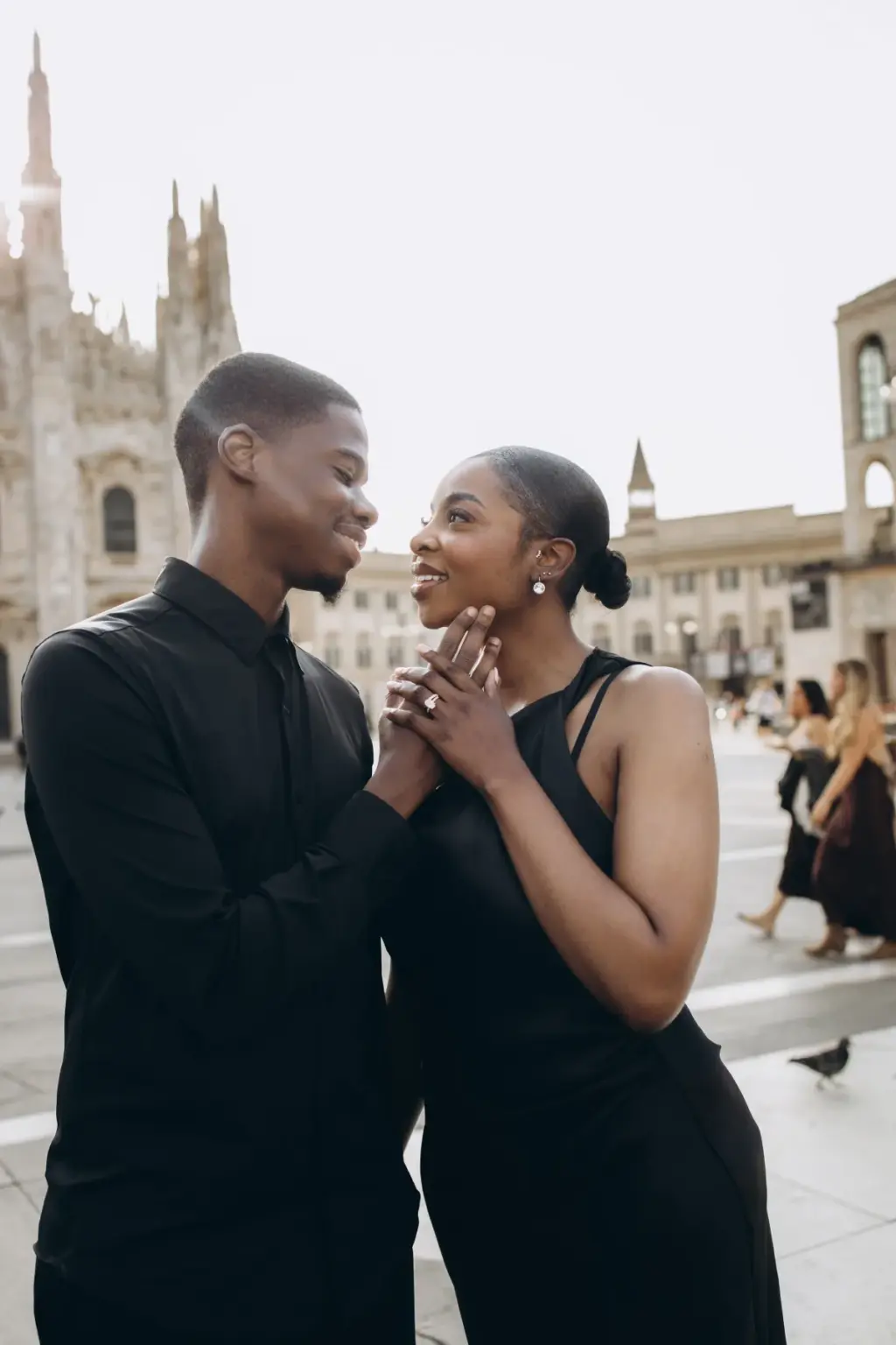 Laughing together, she clasps his hands near her face, ring on show, on the busy square with the Duomo spires to the left.