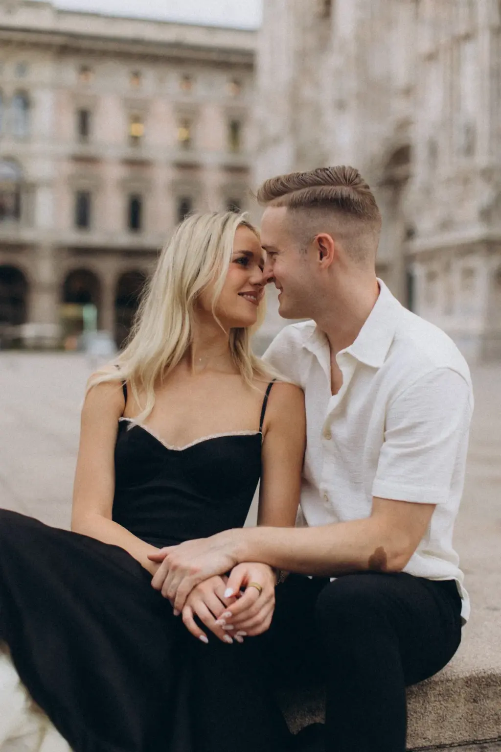 Sitting side by side on a low stone ledge, the couple smiles nose to nose, palazzo facades lining the square behind them.