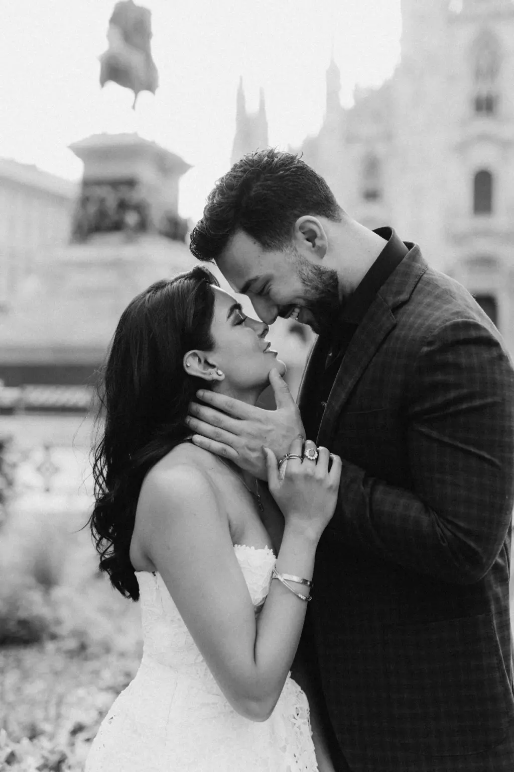 Black-and-white frame of him lifting her chin toward a kiss, the equestrian statue on the Duomo square soft in the background.