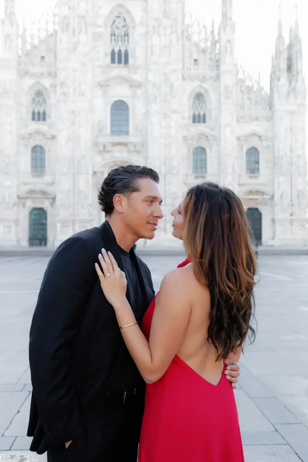 Her ringed hand on his chest, she gazes up at him while he looks off to the side, the Duomo di Milano facade behind them.