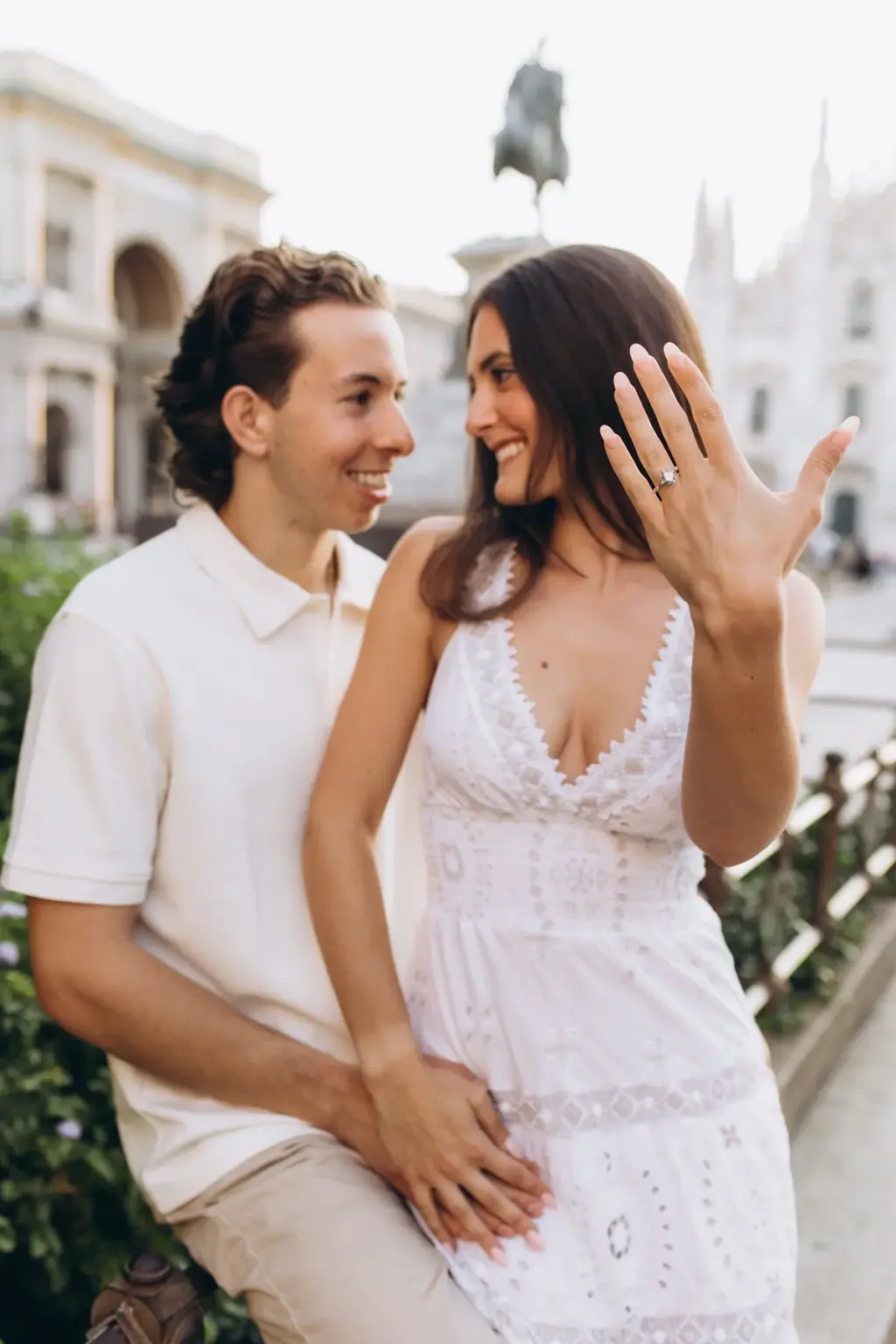 A smiling woman in a white lace dress holds up her hand to show a ring while her partner laughs beside her near the Duomo square.