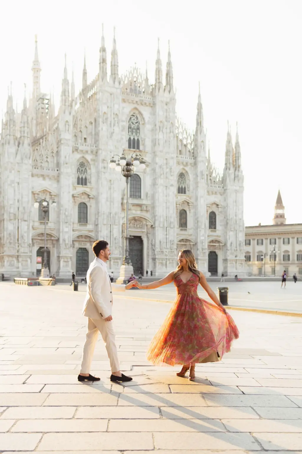 Her pink dress flares as he spins her across the sunlit paving, long shadows stretching toward the Duomo and its tall spires.