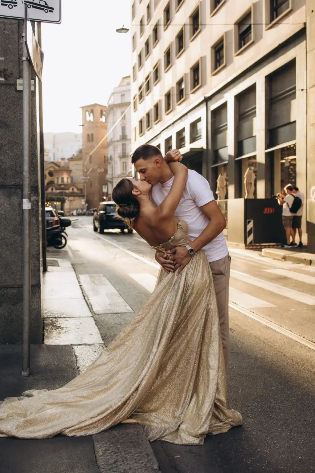 On a Milan crosswalk, a man dips a woman in a gold gown back into a kiss, her train pooled on the pavement around them.