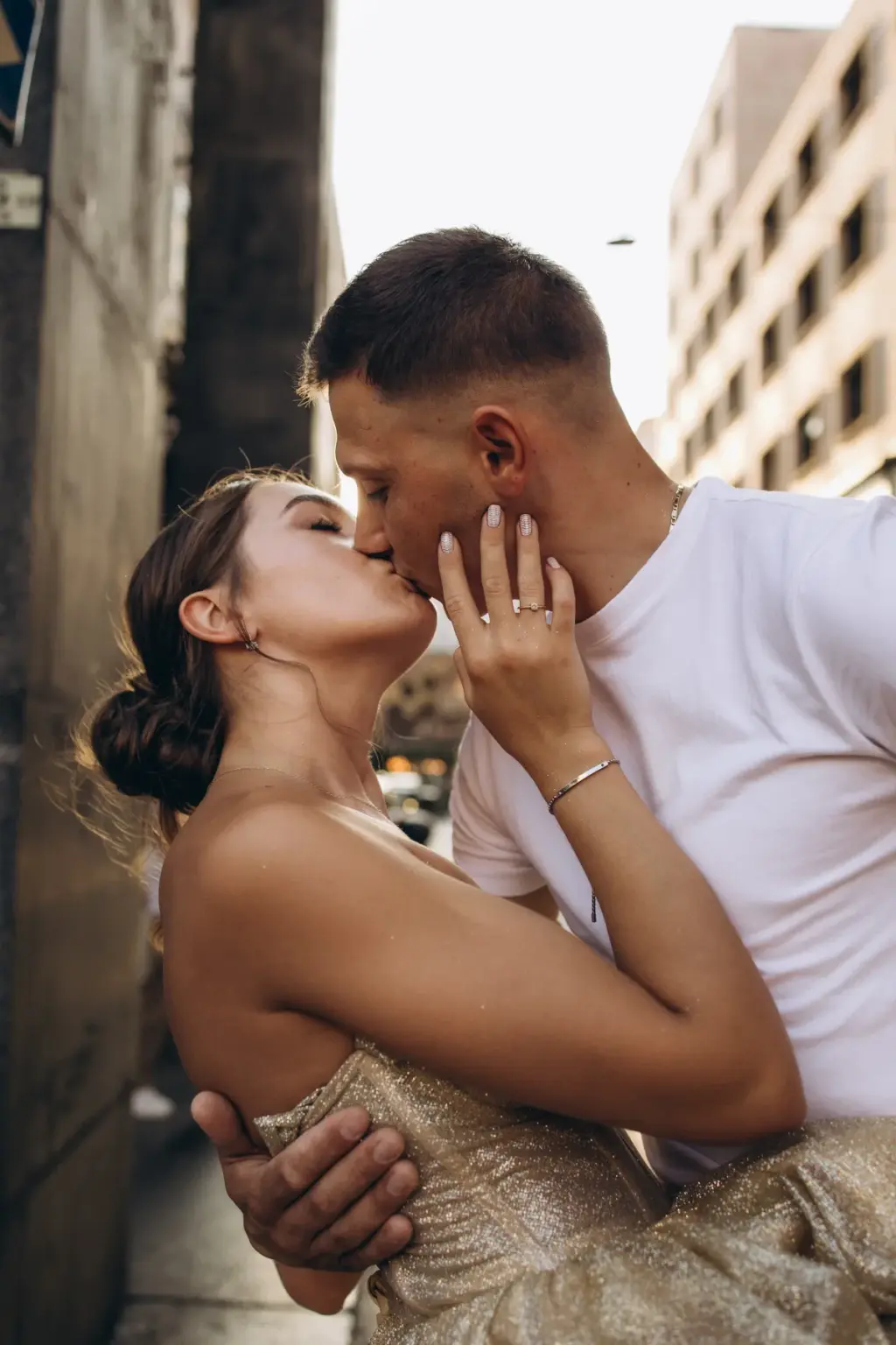 A woman in a gold gown kisses her partner, her hand with a ring resting on his cheek, on a narrow Milan street.