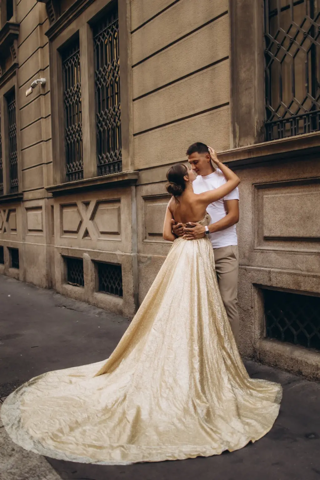 A woman in a gold flying gown with a long sweeping train holds her partner close against a pale stone palazzo on a Milan street.