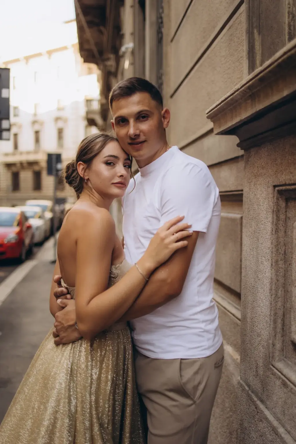 A woman in a sparkling gold strapless gown hugs her partner in a white T-shirt beside a stone building on a Milan side street.
