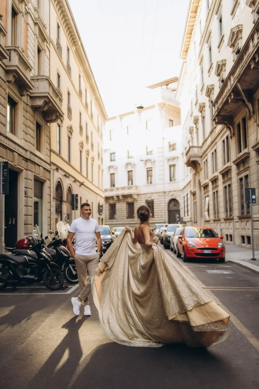 A man in a white T-shirt leads a woman in a voluminous gold gown down the middle of a Milan street lined with parked scooters.