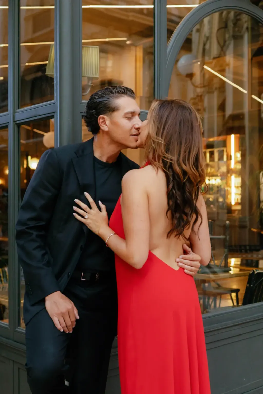She pulls him in for a kiss against the dark-framed windows of a Galleria café, one hand resting on his shoulder.